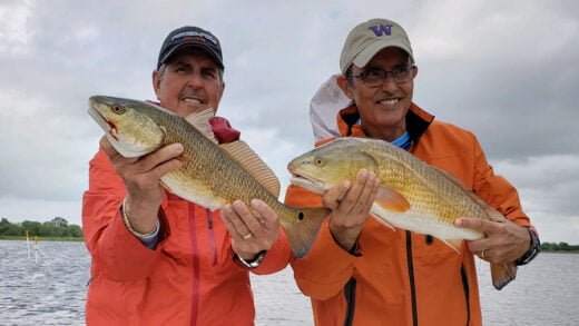 Two smiling anglers holding large Louisiana redfish on a boat with cloudy sky background